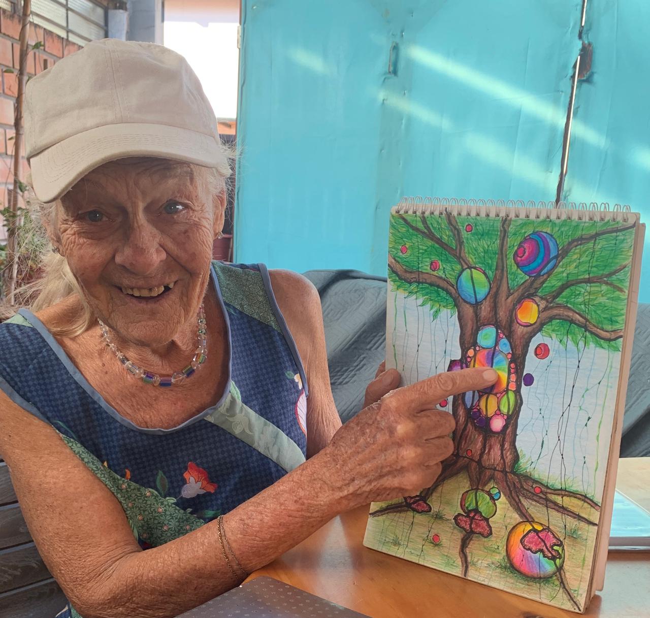 A smiling older woman pointing to her completed colorful neuro art tree drawing in a Microdose Neuro Art class
